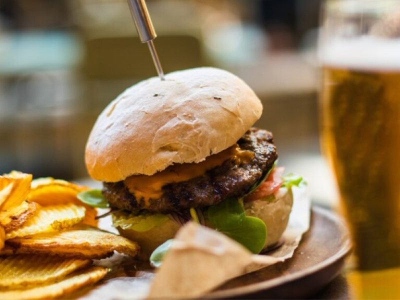 Cheeseburger with lettuce and tomato served alongside crinkle-cut potato chips and a glass of beer.