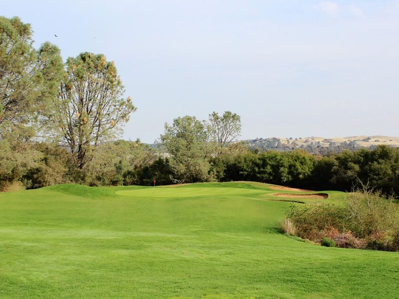 Scenic golf green with a red flag, sand bunker, and rolling Northern California foothills at Castle Oaks Golf Club.