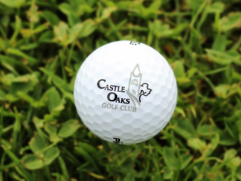 Close-up of a white golf ball featuring the Castle Oaks Golf Club logo resting on green grass.