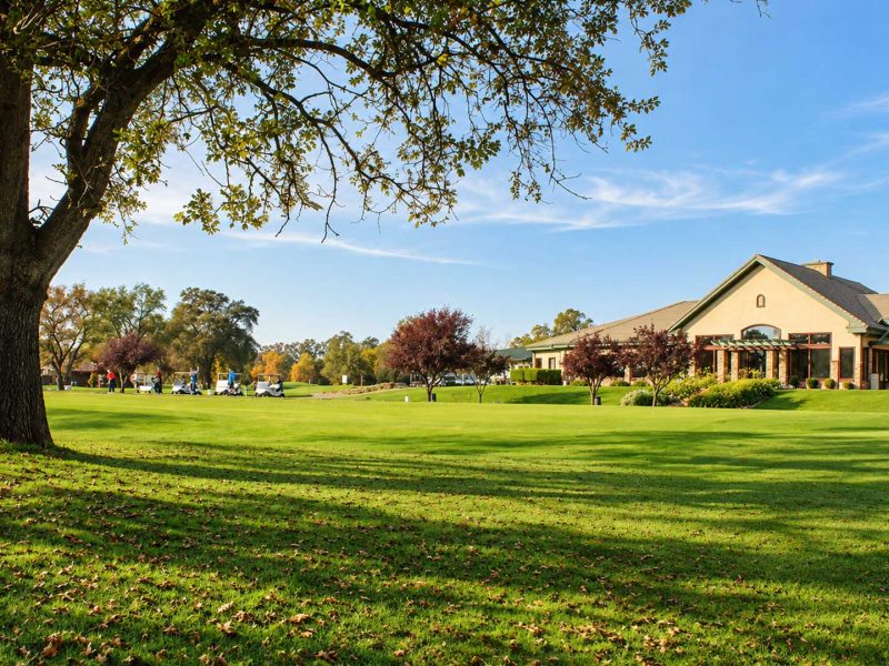 Castle Oaks Golf Club clubhouse and adjacent practice area surrounded by green lawns and mature oak trees in Ione, CA.